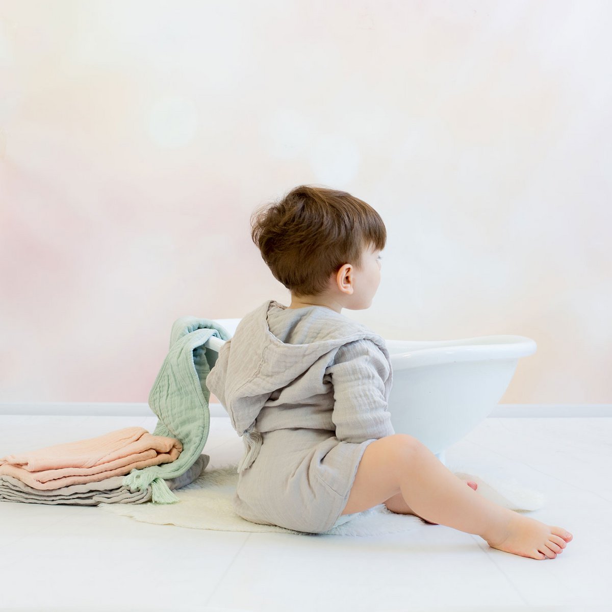 Kleinkind in einem weichen, grauen Musselin-Bademantel mit Gürtel sitzt barfuß vor einer weißen, dekorativen Babybadewanne. Neben der Wanne liegen gefaltete Musselin-Handtücher in Pastellfarben. Der Hintergrund ist sanft und in warmen Beige- und Rosatönen gehalten