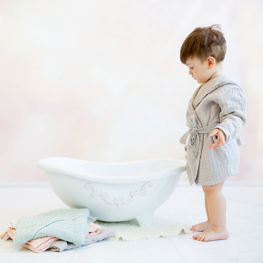 Kleinkind in einem weichen, grauen Musselin-Bademantel mit Gürtel steht barfuß vor einer weißen, dekorativen Babybadewanne. Neben der Wanne liegen gefaltete Musselin-Handtücher in Pastellfarben. Der Hintergrund ist sanft und in warmen Beige- und Rosatönen gehalten