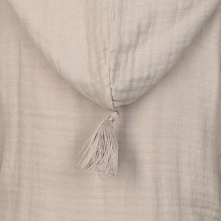 Detailaufnahme einer beigen Musselin-Kapuzenjacke oder eines Bademantels für Kinder. Die Kapuze hat eine dekorative Quaste an der Spitze. Das Material ist weich und leicht strukturiert.