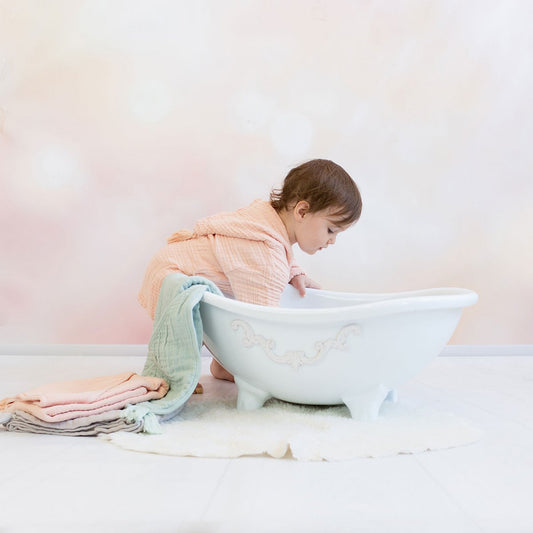Kleinkind in einem weichen, lachsfarbenen Musselin-Bademantel mit Gürtel steht barfuß vor einer weißen, dekorativen Babybadewanne. Neben der Wanne liegen gefaltete Musselin-Handtücher in Pastellfarben. Der Hintergrund ist sanft und in warmen Beige- und Rosatönen gehalten.