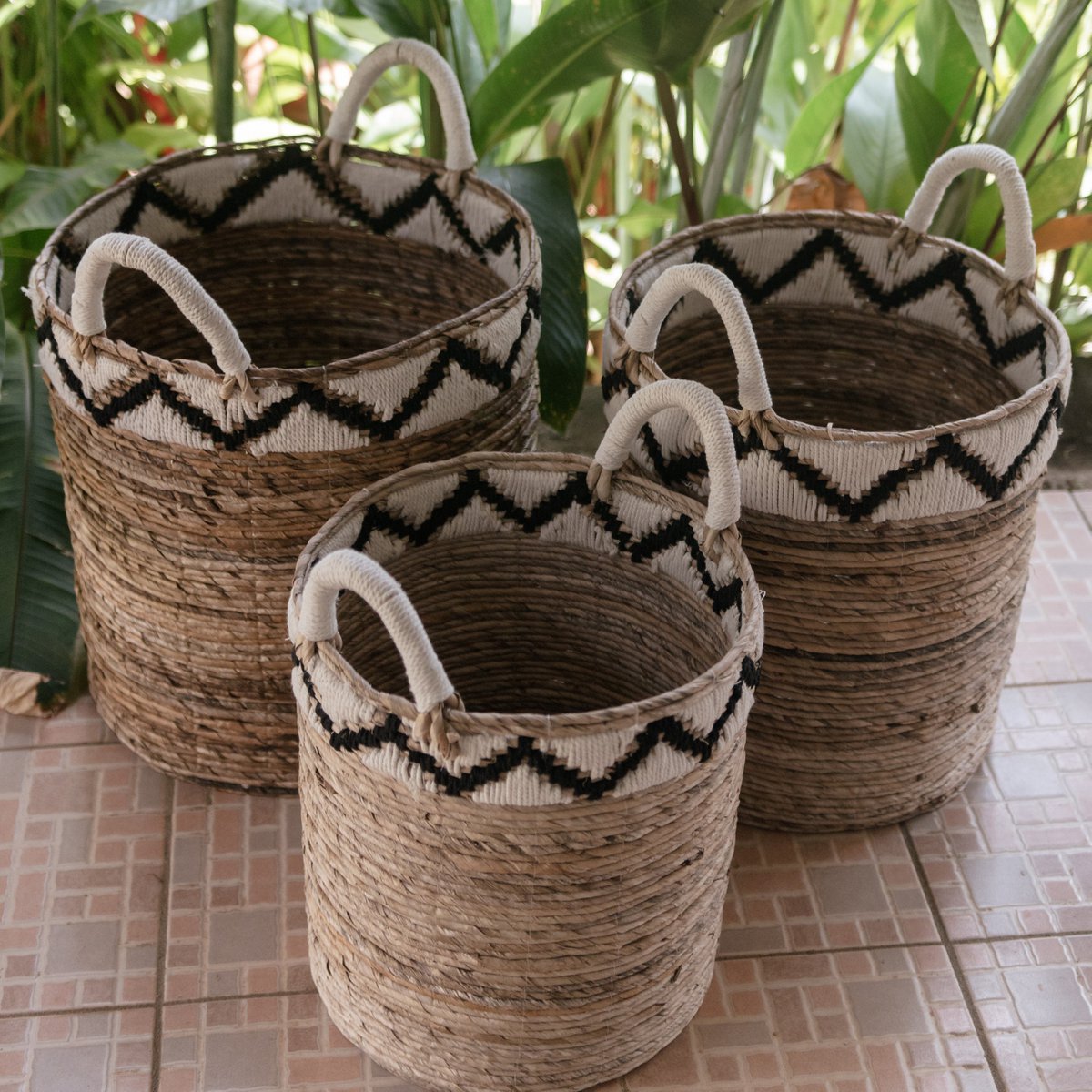 „Korb DALU aus natürlicher Bananenfaser, handgeflochten im Boho-Stil, vielseitig einsetzbar als Wäschekorb, Aufbewahrung oder Pflanzkorb – erhältlich in drei Größen mit attraktivem zickzack Muster am oberen Rand . Die grünen Blätter im Hintergrund bilden einen schönen Kontrast.