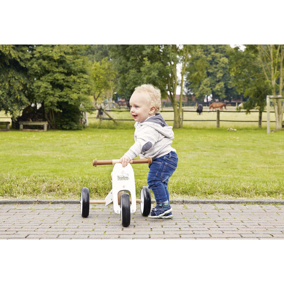 Kind läuft neben Mini-Dreirad 'Charlie' in Weiß und Naturholz, ergonomisches Design, ideal für kleine Kinder ab 1 Jahr