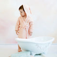 Bild in Galerie-Betrachter laden, Kleinkind in einem weichen, lachsfarbenen Musselin-Bademantel mit Gürtel steht barfuß vor einer weißen, dekorativen Babybadewanne. Neben der Wanne liegen gefaltete Musselin-Handtücher in Pastellfarben. Der Hintergrund ist sanft und in warmen Beige- und Rosatönen gehalten.
