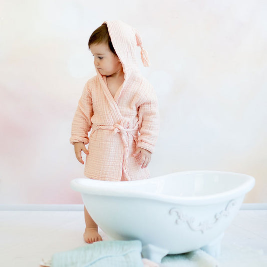 Kleinkind in einem weichen, lachsfarbenen Musselin-Bademantel mit Gürtel steht barfuß vor einer weißen, dekorativen Babybadewanne. Neben der Wanne liegen gefaltete Musselin-Handtücher in Pastellfarben. Der Hintergrund ist sanft und in warmen Beige- und Rosatönen gehalten.