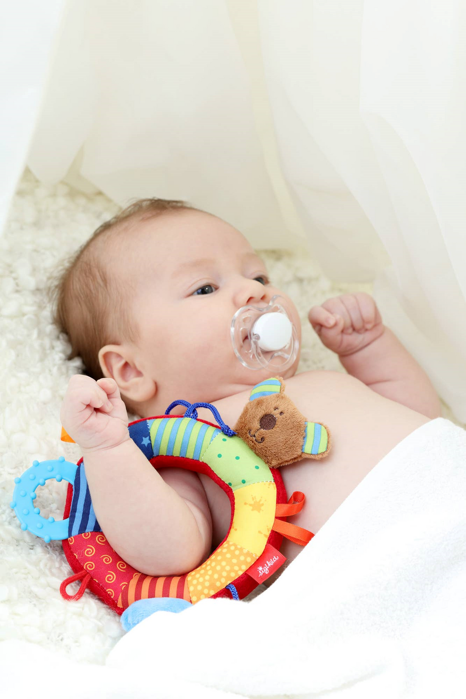 Baby spielt mit Babygreifling Bär, aus buntem Stoff, mit Rassel und Greifring, ideal für kleine Kinderhände.